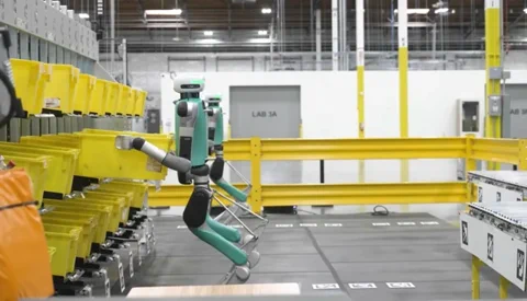 La génération précédente du robot Digit d'Agility, avec ses jambes inversées emblématiques, manipulant des bacs dans un entrepôt.