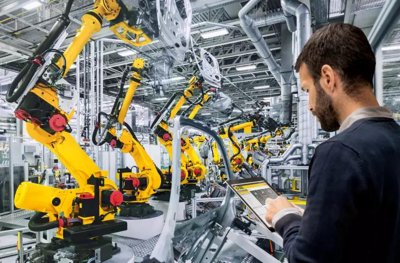 Inženýr používá tablet k řízení tovární haly plné žlutých robotických ramen FANUC.