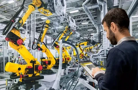 An engineer uses a tablet to manage a factory floor filled with yellow FANUC robotic arms.