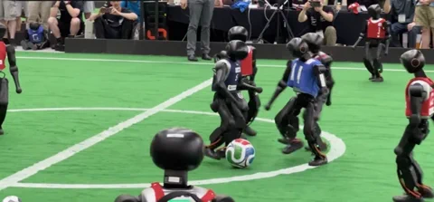 Dva týmy humanoidních robotů hrají fotbal na zeleném hřišti během prvního zápasu 11 proti 11 na RoboCupu 2026.